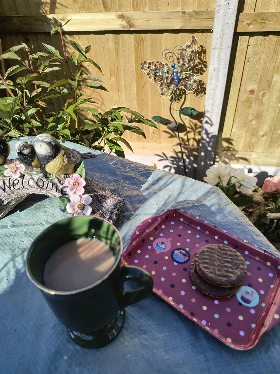 A polka-dot tray with a green mug of tea and chocolate-coated biscuits, sat next to a wooden 'Welcome' garden sign with painted bird and flower ornaments and a butterfly garden ornament against the timber fence, all bathed in afternoon sun.