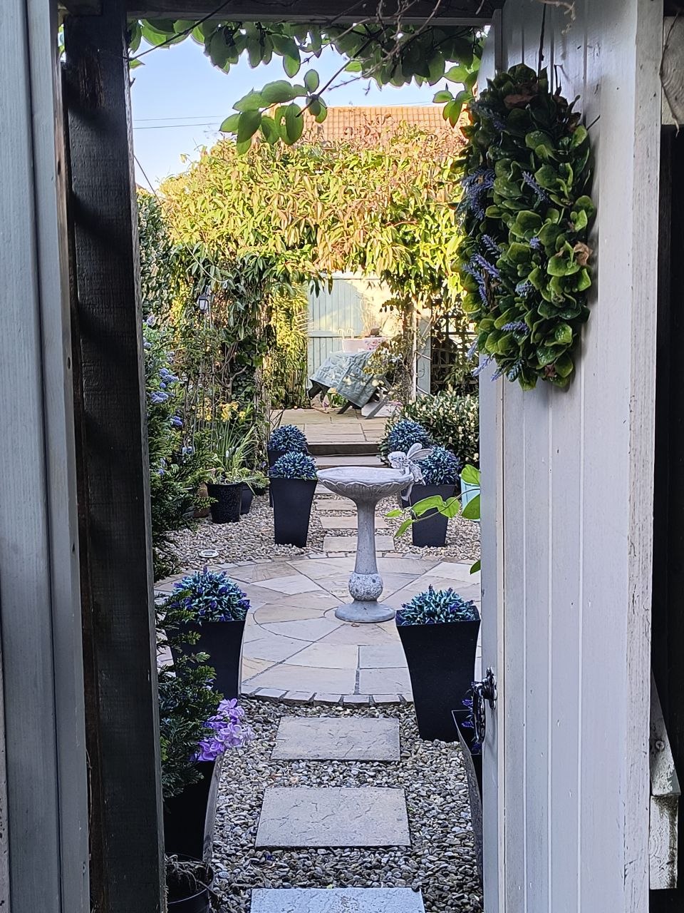 View through a doorway from inside the customer's house out into the courtyard garden in Deal, stone bird-bath in the centre on a paved circle, blue-toned planters with hydrangeas and lavender, gravel-and-stone path, a green leafy wreath hanging on the door frame, end-of-day light.