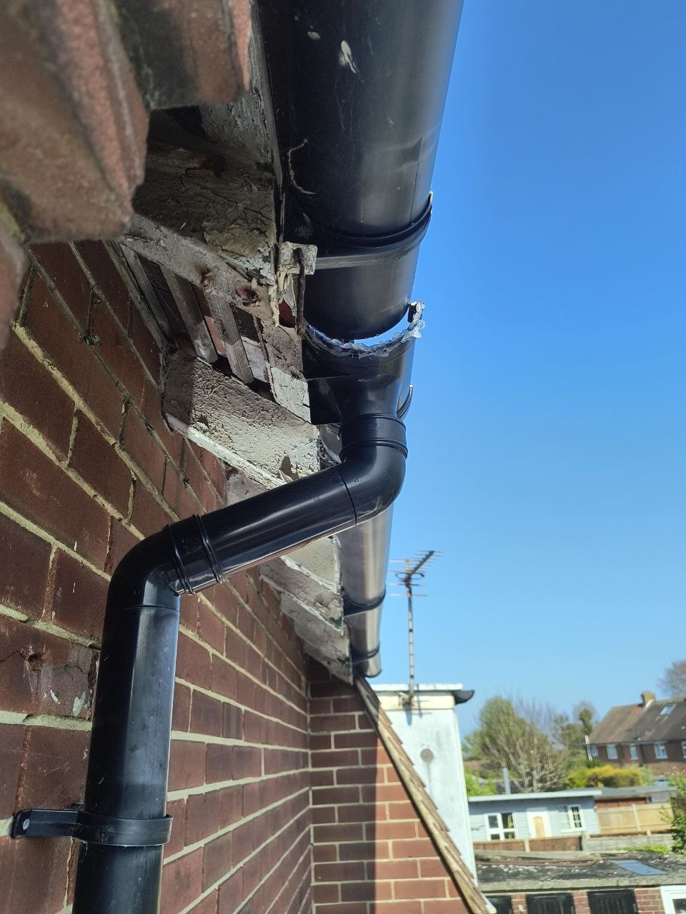 A black plastic gutter and downpipe at the corner of a brick property in Deal, Kent, the gutter joint sitting slightly off, evidence of where water has been spilling onto the wall behind. Bright blue sky behind, neighbouring rooflines visible.