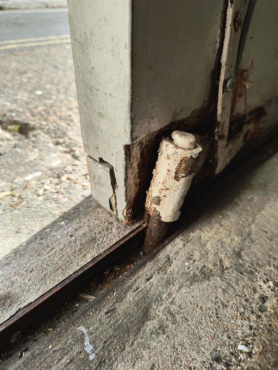 Door frame corner with the lower portion of the door visible, paint and timber damage around the threshold, with a wooden filler peg and rust visible against the sandstone path outside.