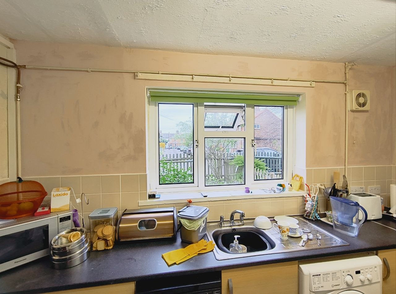 A Folkestone kitchen with a freshly-painted wall in soft Powder Pink, green roller blind in the centre window, sink and worktops below, the room visibly brighter than the patchy peeling wall it replaces.